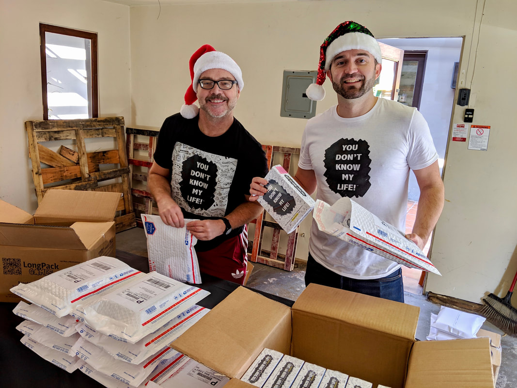 Jeb and Dennis wearing Santa hats and packaging orders of their card game, You Don't Know My Life